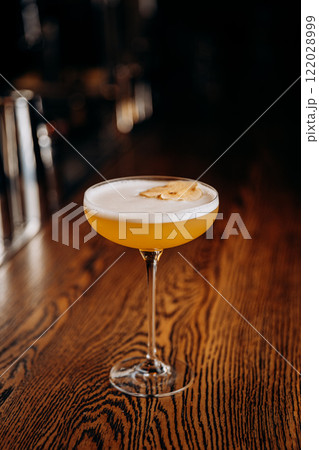 Elegant yellow cocktail with foam and dried apple garnish on a wooden bar top in low light Elegant yellow cocktail with foam and dried apple garnish on a wooden bar top in low light 122028999