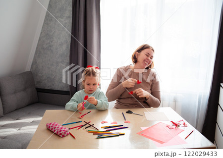 Happy young Caucasian mother teaches her little daughter how to cut hearts out of paper. The concept of Mother's Day and Valentine's Day Happy young Caucasian mother teaches her little daughter how to cut hearts out of paper. The concept of Mother's Day and Valentine's Day 122029433