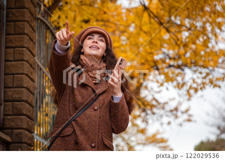 Bottom view of a Caucasian woman in a coat and hat holding a phone, smiling and points forward with finger. City modern lifestyle 122029536