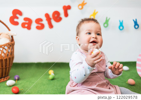 Portrait of a little happy Caucasian girl in a dress, sitting on a green rug with an Easter egg in her hand. In the background, Easter decorations Portrait of a little happy Caucasian girl in a dress, sitting on a green rug with an Easter egg in her hand. In the background, Easter decorations 122029578