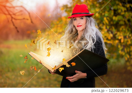 A beautiful woman with a book of magic spells with falling autumn leaves is conjuring 122029673