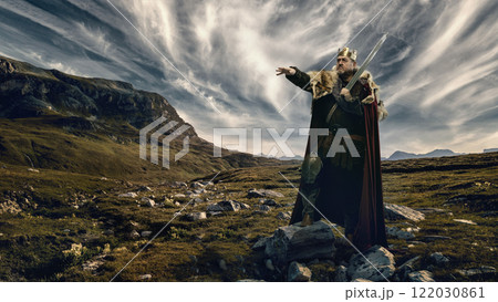Medieval bearded man, king in golden crown, fur-lined cloak, and armor, holding sword and pointing forward, standing on rocky hill with mountains under dynamic cloudy sky Medieval bearded man, king in golden crown, fur-lined cloak, and armor, holding sword and pointing forward, standing on rocky hill with mountains under dynamic cloudy sky 122030861
