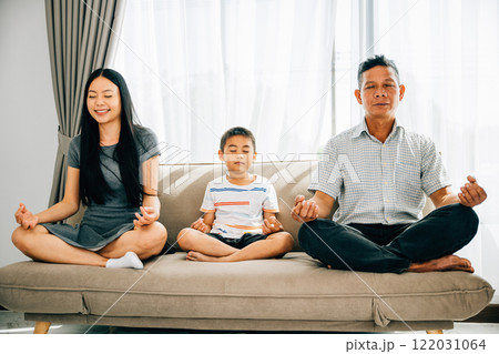 A serene young family practices yoga together seated in lotus position on a sofa. A tranquil moment as parents and their little son meditate fostering harmony and relaxation at home. A serene young family practices yoga together seated in lotus position on a sofa. A tranquil moment as parents and their little son meditate fostering harmony and relaxation at home. 122031064