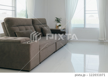 A modern gray sofa with two pillows and built-in drink holders, placed in a bright room with white curtains and floors. Minimalist living room interior with natural light and cozy ambiance. A modern gray sofa with two pillows and built-in drink holders, placed in a bright room with white curtains and floors. Minimalist living room interior with natural light and cozy ambiance. 122031071