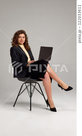 Confident young lady working in laptop, sitting in chair crossing legs against grey studio background. Freelancer. Confident young lady working in laptop, sitting in chair crossing legs against grey studio background. Freelancer. 122033611