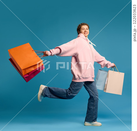 Happy, smiling young woman,wearing casually running in motion with purchases against blue studio background. 122033623