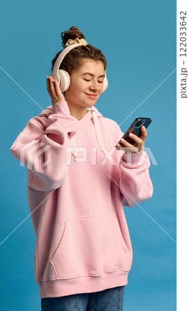 Young, beautiful woman dressed casually, choose song in playlist on phone to listen in in headphones against blue studio background. 122033642