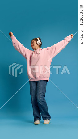 Overjoyed young woman dressed pink hoodie, dancing raising hands while listening music in headphones against blue studio background. Overjoyed young woman dressed pink hoodie, dancing raising hands while listening music in headphones against blue studio background. 122033649