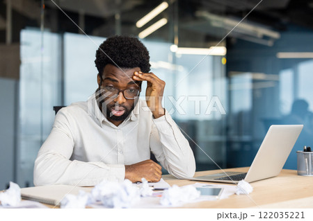 Disappointed and upset man doing paperwork inside office. Unsatisfied with financial reporting results sitting near crumpled pieces of papers. Businessman with laptop among documents. 122035221