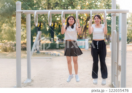 Two women are hanging from a playground swing. One of them is wearing a white shirt and black skirt 122035476