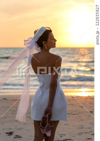 Young elegant woman in white dress and hat on the beach at sunset Young elegant woman in white dress and hat on the beach at sunset 122035827