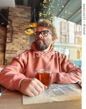 Cheerful man in a peach hoodie smiles warmly while holding a glass of tea. Cozy cafe atmosphere is accentuated by warm string lights and rustic wooden decor, creating a relaxed and inviting vibe in Cheerful man in a peach hoodie smiles warmly while holding a glass of tea. Cozy cafe atmosphere is accentuated by warm string lights and rustic wooden decor, creating a relaxed and inviting vibe in 122035846
