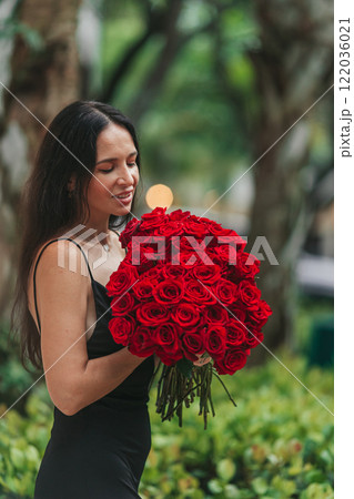 Happy young woman in black dress holding bouquet of red roses outdoors 122036021