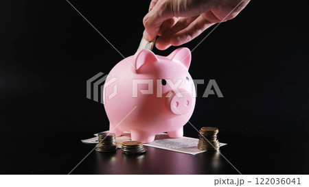 Male hand inserting cash into a pink piggy bank, coins and notes scattered, dark backdrop Male hand inserting cash into a pink piggy bank, coins and notes scattered, dark backdrop 122036041