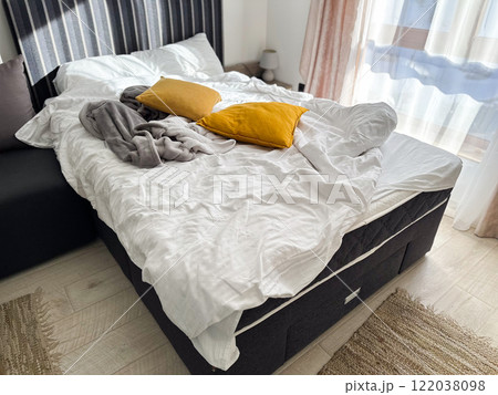 An unmade bed with a white blanket and mustard-yellow pillows in a bright, sunlit room. Mess at home concept 122038098
