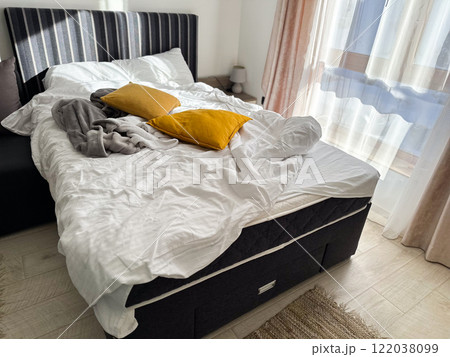 An unmade bed with a white blanket and mustard-yellow pillows in a bright, sunlit room. Mess at home concept An unmade bed with a white blanket and mustard-yellow pillows in a bright, sunlit room. Mess at home concept 122038099