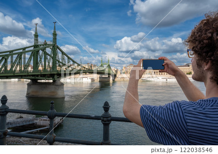 Budapest,Hungary,August 29,2022. A young man takes pictures with his mobile phone of the magnificent cityscape with the Freedom Bridge on a beautiful summer day. Iconic image of the large green bridge 122038546