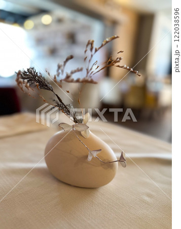 Elegant restaurant table with a small ceramic vase holding dry branches. The blurred background adds depth and highlights the simplicity of the tabletop decor Elegant restaurant table with a small ceramic vase holding dry branches. The blurred background adds depth and highlights the simplicity of the tabletop decor 122038596