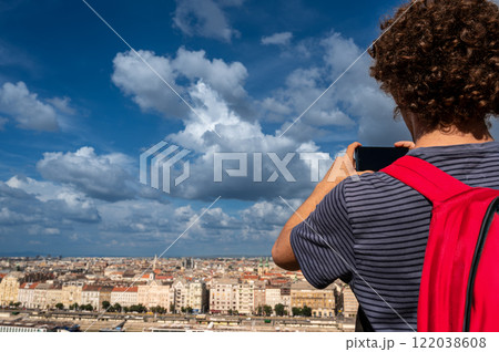 Budapest,Hungary,August 29,2022. A young man takes pictures with his mobile phone of the magnificent cityscape from the top of the hill. Beautiful summer day with blue sky and white clouds. 122038608