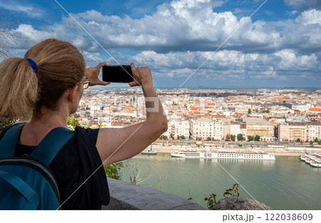 Budapest, Hungary, August 29, 2022. A middle-aged woman takes photos with her mobile phone of the magnificent cityscape from the top of the hill. Beautiful summer day with blue sky and white clouds. Budapest, Hungary, August 29, 2022. A middle-aged woman takes photos with her mobile phone of the magnificent cityscape from the top of the hill. Beautiful summer day with blue sky and white clouds. 122038609