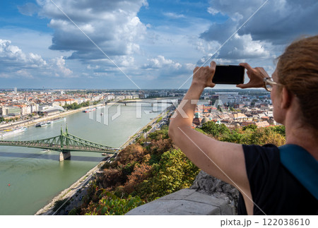 Budapest, Hungary, August 29, 2022. A middle-aged woman takes photos with her mobile phone of the magnificent cityscape from the top of the hill. Beautiful summer day with blue sky and white clouds. 122038610