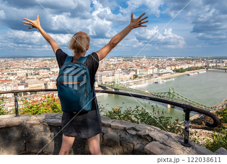Budapest,Hungary,August 29,2022. A middle-aged woman is inspired by the magnificent cityscape from the top of the hill, raises her arms to the sky. Beautiful summer day with blue sky and white clouds. 122038611