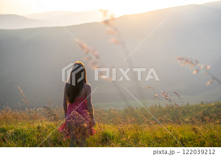 Young woman in red dress standing on grassy field on a windy evening in autumn mountains enjoying view of nature. Young woman in red dress standing on grassy field on a windy evening in autumn mountains enjoying view of nature. 122039212