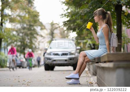 Young woman drinking coffee from paper cup sitting on city street bench in summer park. Young woman drinking coffee from paper cup sitting on city street bench in summer park. 122039222