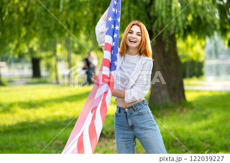Young red haired woman holding USA national flag standing outdoors in summer park. Positive girl celebrating United States independence day. Young red haired woman holding USA national flag standing outdoors in summer park. Positive girl celebrating United States independence day. 122039227