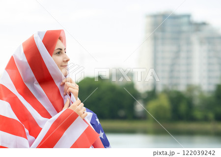 Young happy refugee woman with USA national flag on head and shoulders. Positive muslim girl traveling in United States. 122039242