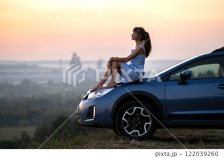 Yong happy female driver resting on hood of her car enjoying sunset view of summer nature. Travel destinations and recreation concept. 122039260