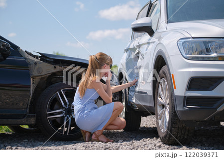 Sad young woman driver sitting near her smashed car looking shocked on crashed vehicles in road accident 122039371