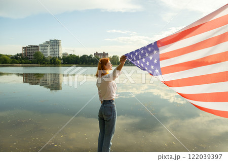 Red haired girl holding USA national flag in her hands. Positive young woman celebrating United States independence day. Red haired girl holding USA national flag in her hands. Positive young woman celebrating United States independence day. 122039397