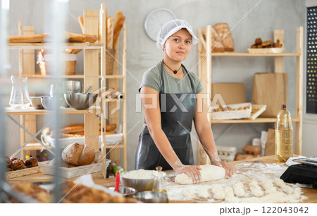 Young baker girl is standing near work table in kitchen, working with dough 122043042
