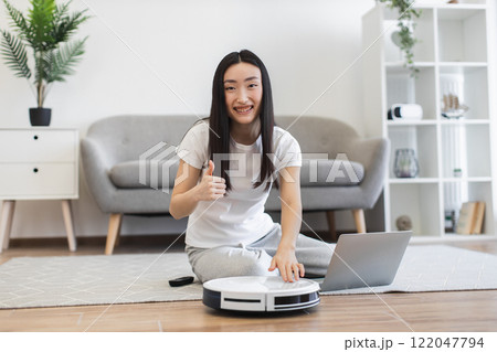 Asian young woman with robot vacuum cleaner on hardwood floor. She smiles and gives thumbs up. Concept of modern technology and easy cleaning. 122047794