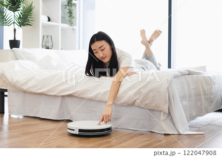 Asian woman relaxing on bed while controlling robot vacuum cleaner. Concept of modern technology, convenience, smart home lifestyle. Young adult enjoying clean and tidy living space. Asian woman relaxing on bed while controlling robot vacuum cleaner. Concept of modern technology, convenience, smart home lifestyle. Young adult enjoying clean and tidy living space. 122047908