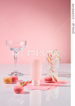 An advertising product photo with a close-up angle. Party table theme featuring macarons, candy canes in a glass, and a pink cosmetic bottle mockup on a white surface against a pink backdrop. 122049144