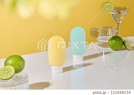 Side angle shot of two squeeze bottles positioned centrally on a white surface. Limes, a martini glass, and a glass pitcher arranged against a yellow background. The fruit theme for cosmetics ads. 122049154