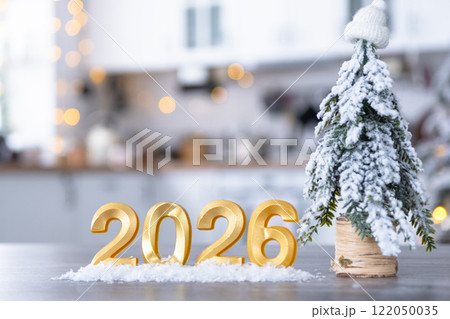 Golden number 2026 on table in background of cozy white kitchen with Christmas tree and Christmas decor with garland lights. Housing, interior, renovation, mortgage, rent in New Year 122050035