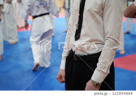 A close-up of a row of young athletes in white karate kimonos with clenched fists and the referee is in the foreground. 122054984