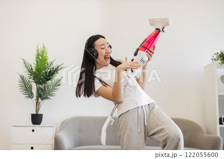Asian woman in her twenties enjoying music with headphones while vacuuming. Captures fun and playful cleaning at home, conveying energy and happiness during household chores. Asian woman in her twenties enjoying music with headphones while vacuuming. Captures fun and playful cleaning at home, conveying energy and happiness during household chores. 122055600