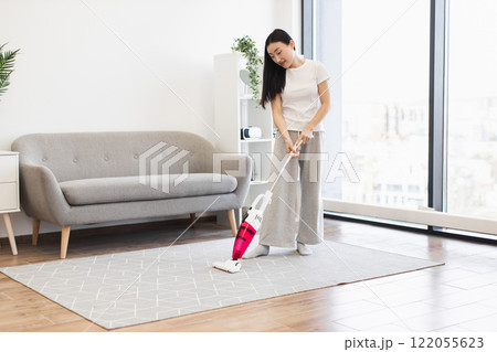 Young Asian woman cleaning living room with vacuum cleaner. Modern bright interior with sofa and plants. Concept of home cleanliness and domestic chores. Young Asian woman cleaning living room with vacuum cleaner. Modern bright interior with sofa and plants. Concept of home cleanliness and domestic chores. 122055623