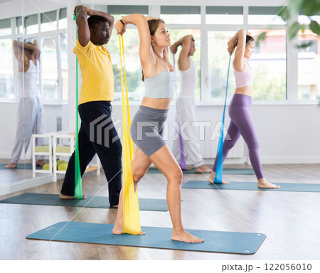 Girl conducts Pilates training for multinational students, standing exercises with wide elastic band Girl conducts Pilates training for multinational students, standing exercises with wide elastic band 122056010