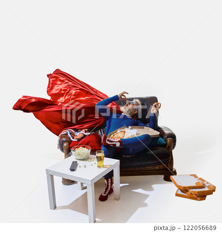 Relaxed man in superhero costume and 3D glasses lounging on couch, eating pizza, table with beer glass and popcorn standing near 122056689
