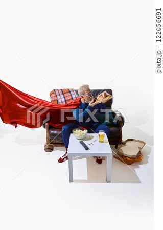 Grey-haired man in superhero costume sitting on couch, eating pizza, table with beer glass and popcorn standing near, against white studio background 122056691