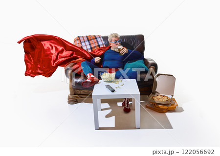 Man in superhero costume and 3D glasses sitting on couch, eating popcorn and sipping beer, open pizza boxes on floor against white background 122056692