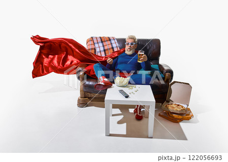 Man in superhero costume and 3D glasses sitting on couch, watching TV, eating popcorn and drinking beer, open pizza boxes on floor against white background Man in superhero costume and 3D glasses sitting on couch, watching TV, eating popcorn and drinking beer, open pizza boxes on floor against white background 122056693