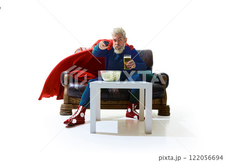 Evening chill at home, Man in superhero costume sitting on couch with remote TV controller, drinking beer, table filled with popcorn against white studio background 122056694