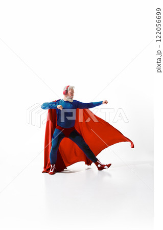 Dynamic image of grey-haired man superhero costume with red cape, listening to music in headphones and standing with fist up against white studio background 122056699
