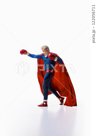 Dynamic image of man in superhero costume with red cape delivering powerful punch in boxing gloves against white studio background Dynamic image of man in superhero costume with red cape delivering powerful punch in boxing gloves against white studio background 122056711